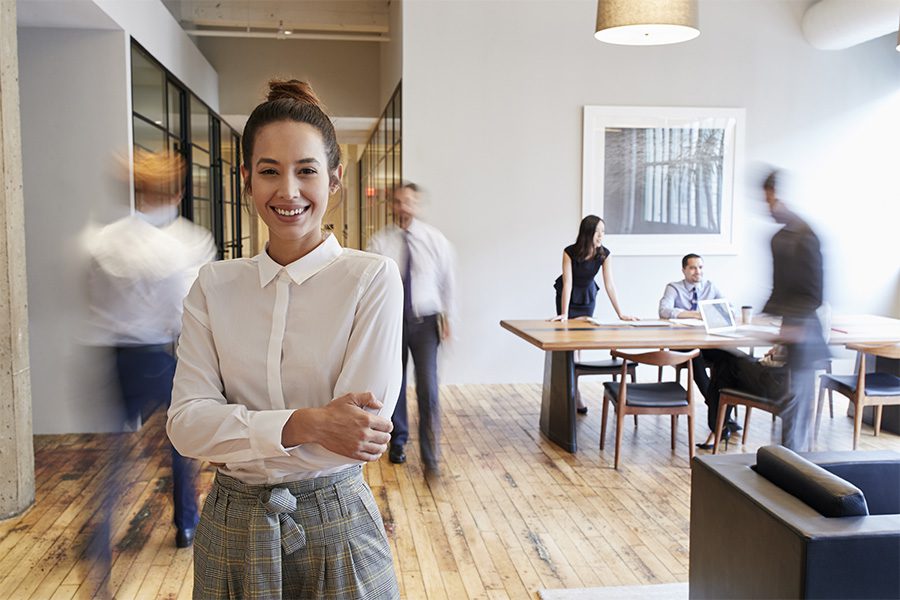 Business Insurance - Portrait of Young Woman Standing in Front of a Busy Modern Workplace Blurred in the Distance