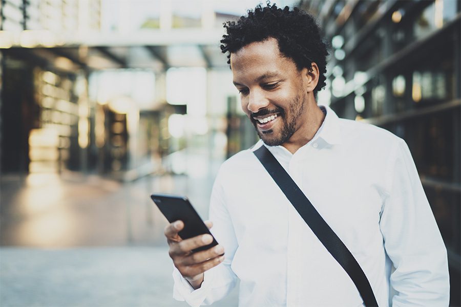 Client Center - Smiling Man Walking in an Ohio City Holding His Phone in His Hands and Checking His Email with the Background Blurred